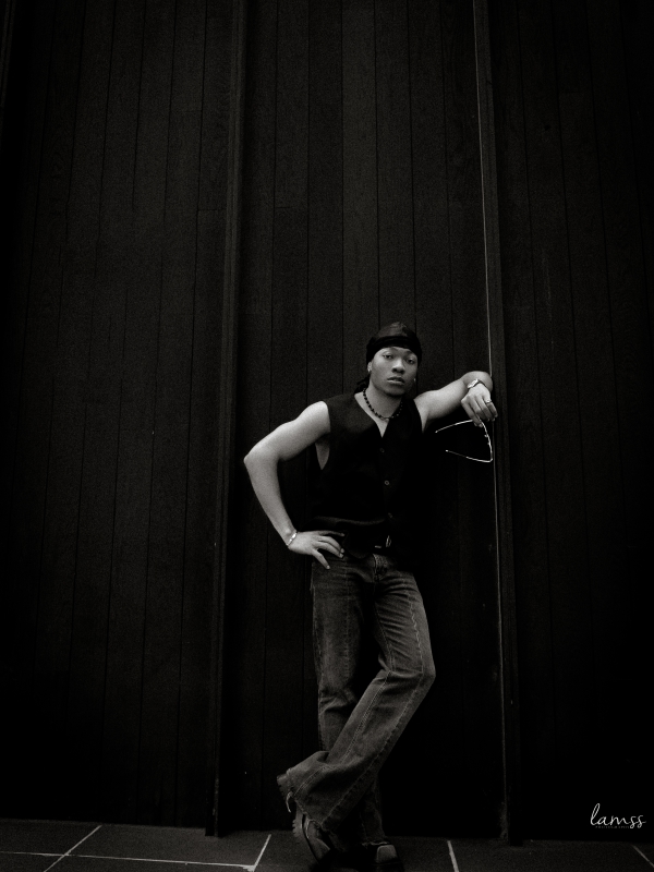 Black And White Full Body Shot Of A Person In A Vest And Jeans Leaning Against A Dark Textured Wall.